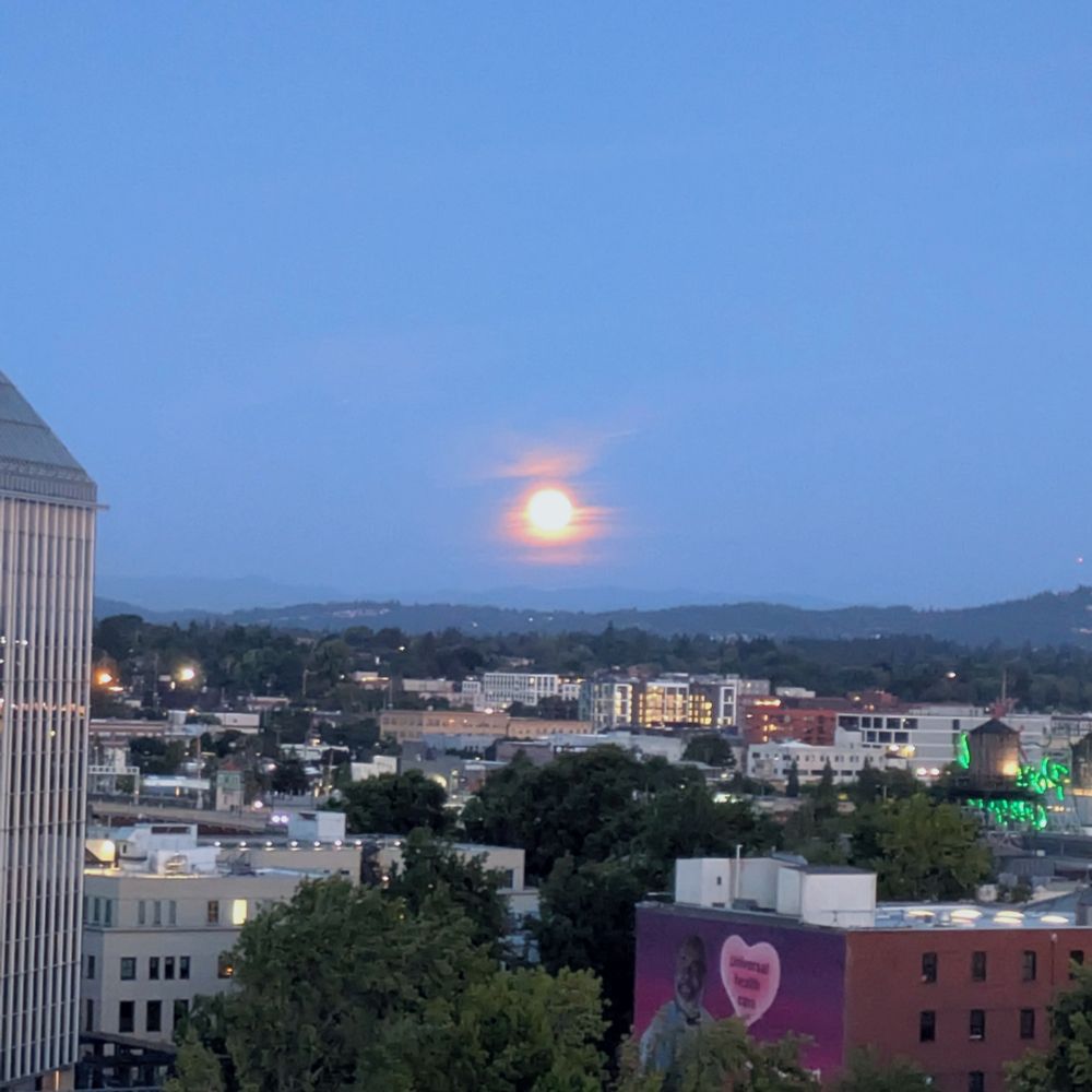 A picture of the moon rising over the hills of a city. Various buildings lay in between