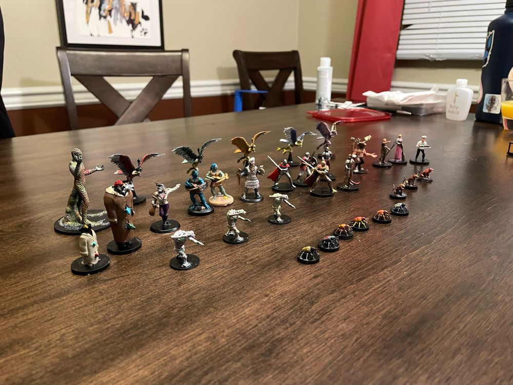 An assortment of completed miniatures displayed among a table. The minis include: Harpies, city guards, commoners, skeleton owlbears, and beetles.