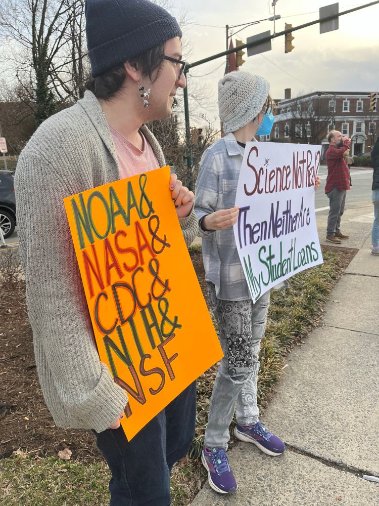 Two rally-goers hold signs at the Charlottesville Stand Up for science event. One sign says “NOAA and NASA and CDC and NIH and NSF.” The other says “Science not real? Then neither are my student loans.”