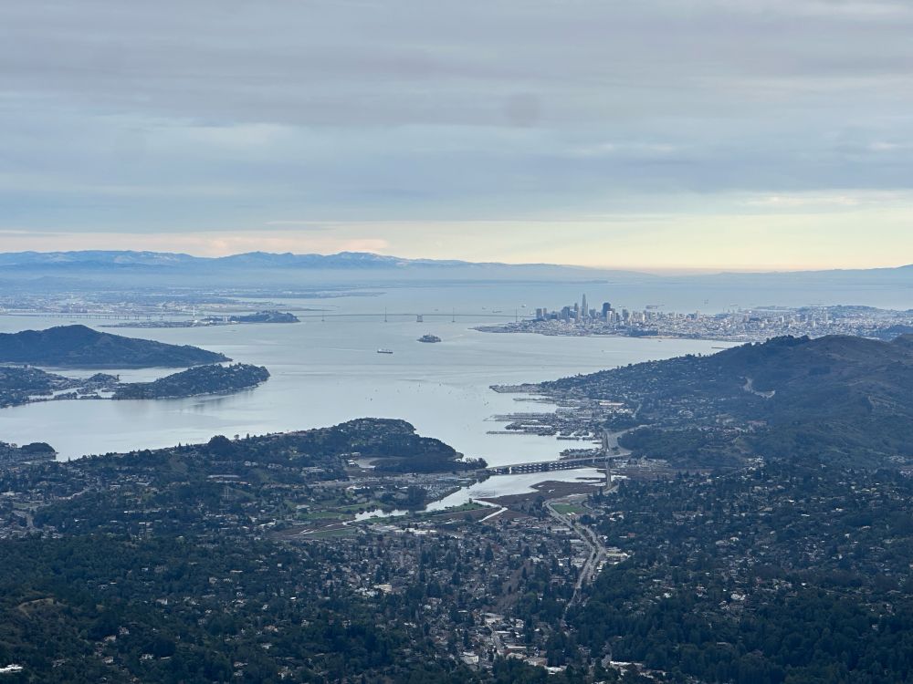 A distant view of cityscapes and the San Francisco Bay from the top of Mount Tamalpais. The bay bridge is visible, and San Francisco and Oakland are recognizable, if tiny.