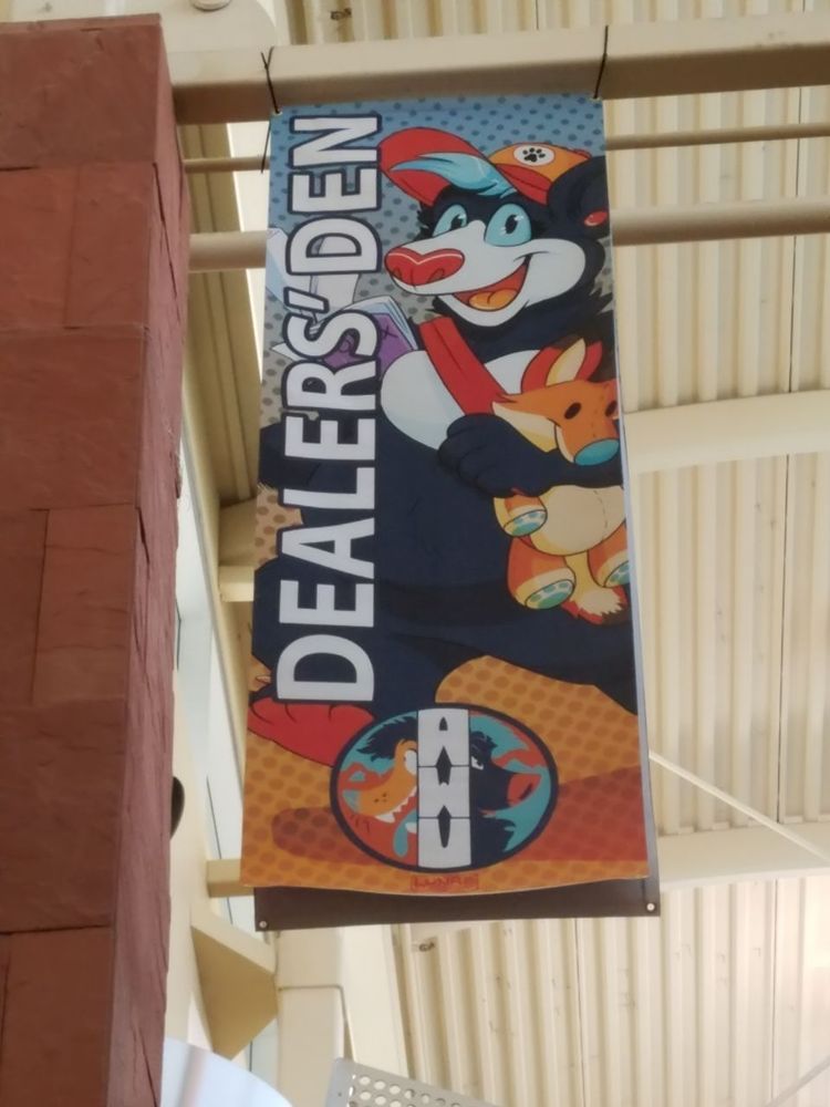 A large vinyl banner hanging from the ceiling of a conference center.

A blue cartoon bear (Carver) is holding a Plush Coyote (Arc), a rolled up poster, a comic book, and is wearing a Paw print hat

'DEALERS' DEN' is listed in large letters