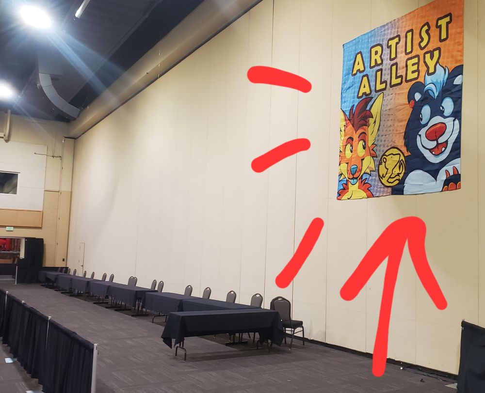 Photo of a conference room.
Several tables are setup along a panel wall.

A large 'Artist Alley' 6 x 6 foot banner hangs on the wall featuring the mascots of AWU, Arc and Carver


