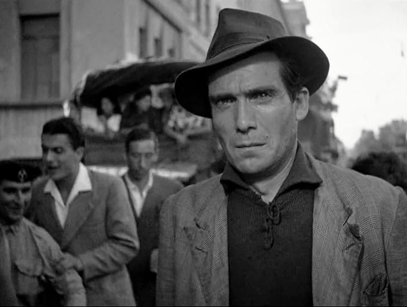 A black-and-white still from the film ‘Bicycle Thieves,’ featuring a close-up of a man wearing a worn jacket and a dark fedora. His expression is tense and somber, reflecting the emotional struggles of the film. In the blurred background, a bustling street scene includes other men in suits and a cart, capturing the post-war Italian urban environment.