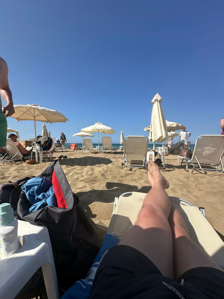POV auf einer Liege am Strand liegend, im Vordergrund andere liegen und Schirme, im Hintergrund (30m) das Mittelmeer. Strahlend blauer Himmel.