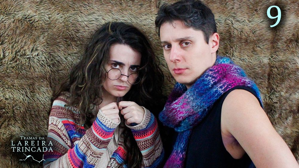 Bruna e Lucas, lado a lado. Bruna está encolhida à esquerda, com as mãos perto do pescoço, como se segurasse algo pequeno. Ela olha por ciam dos óculos redondos fixamente para a câmera, preocupada. Ela veste um casaco marrom com algumas listras azuis e vermelhas. Lucas, à direita, está meio de lado, olhando sério para a câmera com as mãos na cintura. Ele usa uma regata preta e um cachecol azul e roxo.
O fundo atrás é como a pele de um animal estendida, marrom.