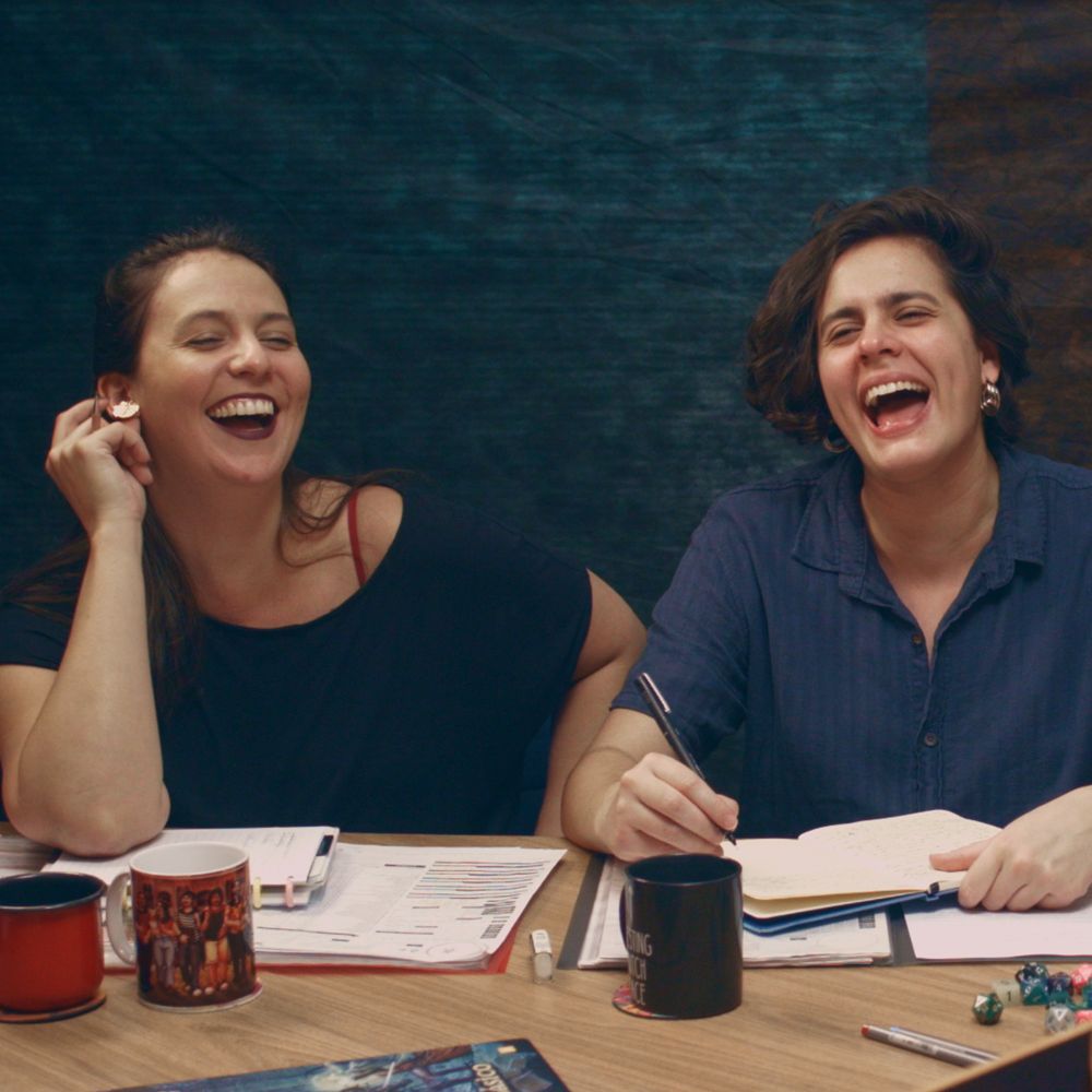 Gabi e Fai, sentados lado a lado em uma mesa. Gabi está rindo enquando segura o brinco em sua orelha direita. Ela usa uma blusa preta. Fai, no meio de escrever algo em seu caderno, também está rindo loucamente. Ele veste uma camisa azul-marinho. A mesa está cheia de coisas de RPG: fichas, dados, canecas. O fundo é um belo fundo de estúdio feito de pano e luz.
