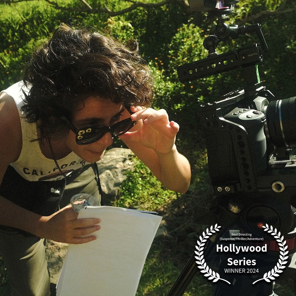 Fai, bem de perto. À câmera está à direita, e Fai está abaixado para olhar para sua tela. Ele segura os óculos escuros com a mão esquerda e olha por cima deles. Com a mão direita, segura um copo d'água e um roteiro. O fundo é uma mata bem verde ao sol. No canto há um louro do prêmio, dizendo: Best Directing (Suspense/Thriller/Adventure) HOLLYWOOD SERIES WINNER 2024