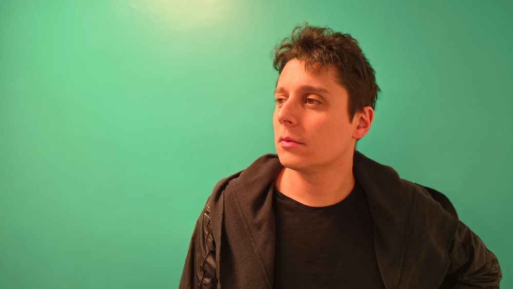 Lucas, em plano médio, olhando meio de canto de olho para o horizonte à esquerda do quadro. Ele usa uma jaqueta preta com uma camiseta preta por baixo. Sua expressão é solene. Atrás dele, o fundo é verde água.