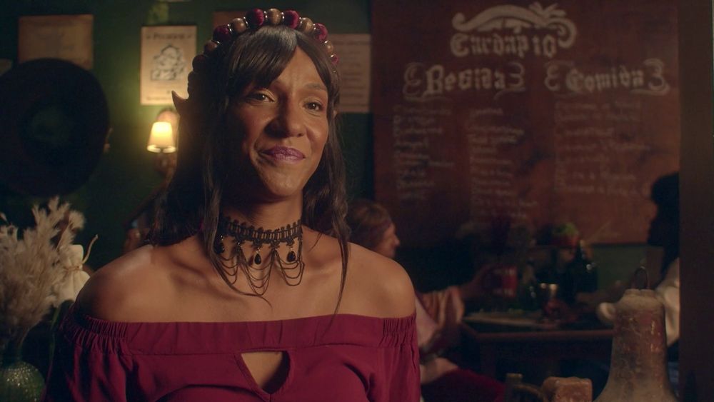 Pedrita, a elfa maga/estagiária em plano médio. Ela veste uma blusa vermelha que deixa seus ombros de fora, e tem uma gargantilha preta elaborada, além de um adorno na cabeça de contas vermelhas e douradas. Atrás dela há o fundo desfocado da Taverna Original, com seu cardápio na parede, paredes verdes e cartazes de procurado. Vig está sentado em uma mesa bem, atrás dela, segurando uma caneca. 
Pedrita é interpretada por Danna, uma mulher trans negra com olhos castanhos e cabelos castanhos na altura dos ombros.