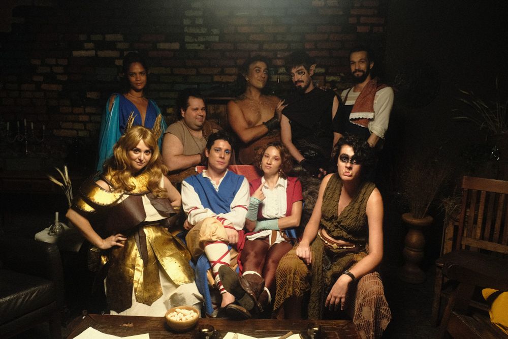 Elenco da websérie Desaventureiros sorrindo para a foto no cenário da Outra Taverna. No sofá: Shura, a anã paladina, Vic, o bardo humano, Vicky, a barda humana, Geburath, a clériga 1/8 orc. Atrás deles, de pé: Shi, a elfa feiticeira, Touro Vegano, o anão guerreiro, Salaz, o meio-elfo druida, Fliq, o meio-elfo ladino, e Enzo, o taverneiro.