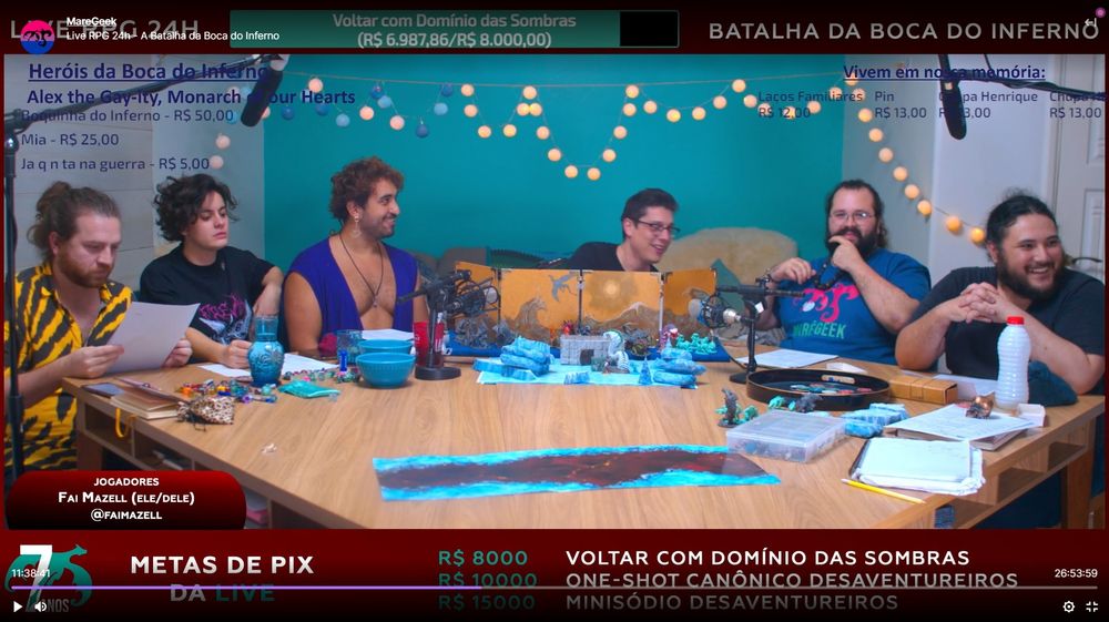 Um screenshot da live: Lucas no centro da mesa, atrás de seu escudo de mestre, rindo. Aran, Fai e Seh à esquerda, Tuzão e Yurik à direita. Sobre a mesa, um caos de dados, potinhos e mapas.