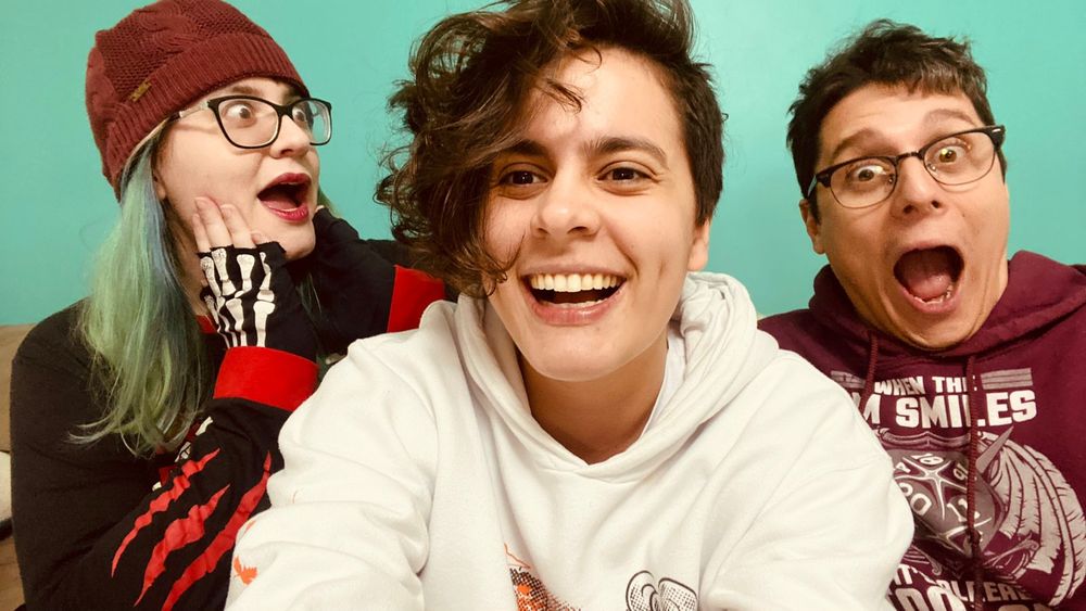 Selfie de Thais, Fai e Lucas contra a parede verde-água da Maré Geek. Thais e Lucas estão boquiabertos e de olhos arregalados, ela olhando para o lado, ele para a frente. Fai olha para a câmera, dando risada.