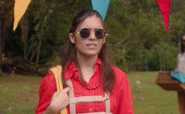 Juni, um humano não-binário, em primeiro plano no dia do expurgo. Ele usa uma blusa vermelho-fiscal, óculos escuros redondos de metal e um colar de contas vermelho. Por cima de tudo, um arnês marrom-claro. No ombro direito ele tem uma bolsa amarela de tecido que segura com a mão direita entuanto olha, transtornado, para algo fora de quadro. Atrás dele, bandeirinha do expurgo em amarelo, azul e vermelho, um piquenique sendo montado e uma grande floresta ao fundo.
Juni é interpretado por Lenny,  pessoa agênero de pele branca, cabelos castanhos longos.