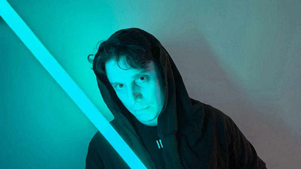 


Lucas, em plano médio. ELe usa uma capa preta com capuz, e olha para a câmera sério. Seu roso é iluminado pela luz azulada de uma espada de luz azul!
