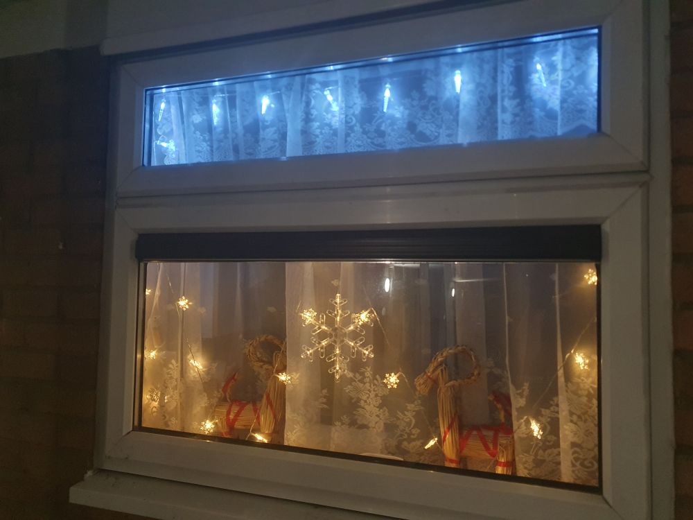 Some Christmas decorations in a window. The row of white lights at the top are icicles and the yellow lights are snowflakes  with a larger snowflake in the middle. There is also two Yule goats facing each other on the window sill. There is a net curtain behind the lights for privacy.