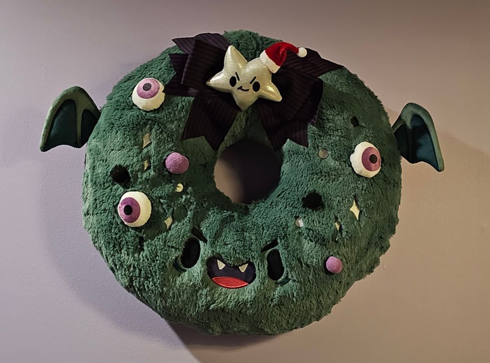Photo of a plush wreath, that is monster themed. It's fuzzy green; decorated with little wings, eyeballs, and an angry little star wearing a Santa hat
