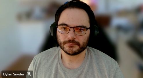 A picture of a 32 year old white male with a beard, wearing headphones, a grey shirt and a new pair of blue light blocking glasses. The background is blurred as he is in a Teams call.