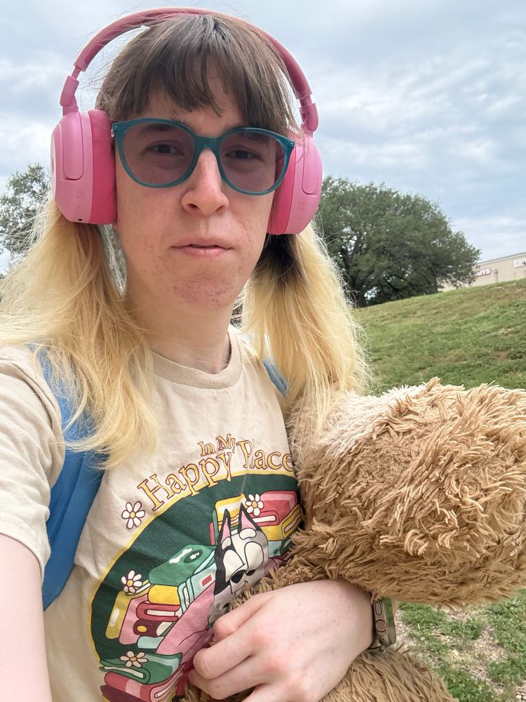A girl with long blonde and brown hair is wearing pink over-ear headphones, teal glasses, and a beige T-shirt with colorful books and ‘Muffin’ from the show ‘Bluey’ that says “In My Happy Place.” They are holding a large, fluffy teddy bear and standing outside near a grassy hill under a cloudy sky.