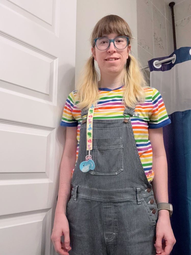 A girl stands in a bathroom wearing a colorful rainbow-striped t-shirt and pinstriped overalls with button details on the side. They have shoulder-length blonde hair in pigtails, blue glasses, and a pastel-colored pacifier clipped to their overalls. A rainbow shower curtain and a white paneled door are visible in the background.