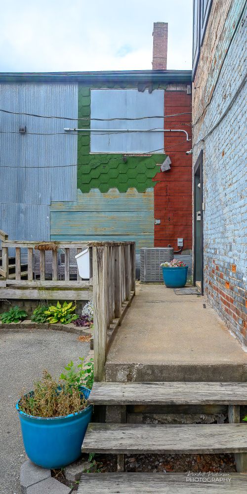 View of a wooden stairs and porch leading to an entrance door.  The wall in the background of the image is covered in multiple types of siding including red brick looking asphalt shingles, Green hexagon shaped asphalt shingles, corrugated metal sheeting and smooth metal sheeting.