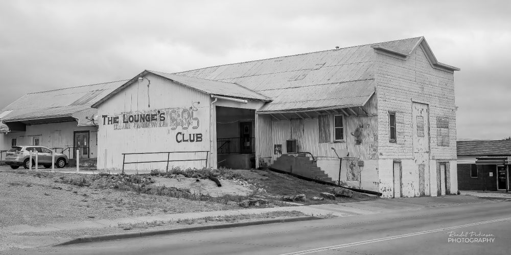 Black and white view of an old commercial building with a drive-through garage on the side that is now used as a bar.  "The Lounge's 5:05 Club" is painted on the side of the building.