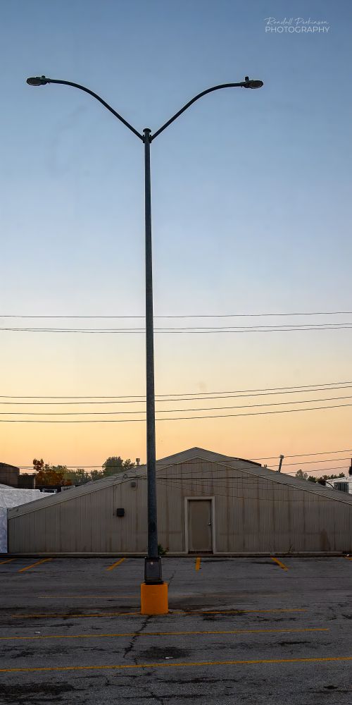 A view of a one story metal building with a peaked roof and one door and its parking lot at dawn.  One parking lot light pole on a yellow concrete base  supports two lights.