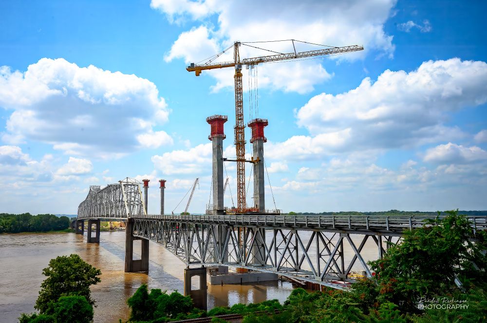 A view from the Illinois side of the Chester Bridge which is a steel truss bridge built in the 1940s that includes 2 660' long center spans.  Adjacent to the old bridge you see two new bridge towers being constructed, one with a tower crane in position between the tower legs.  A third tower is located beyond these two at the Missouri side of the river that can't be seen due to the truss of the old bridge.