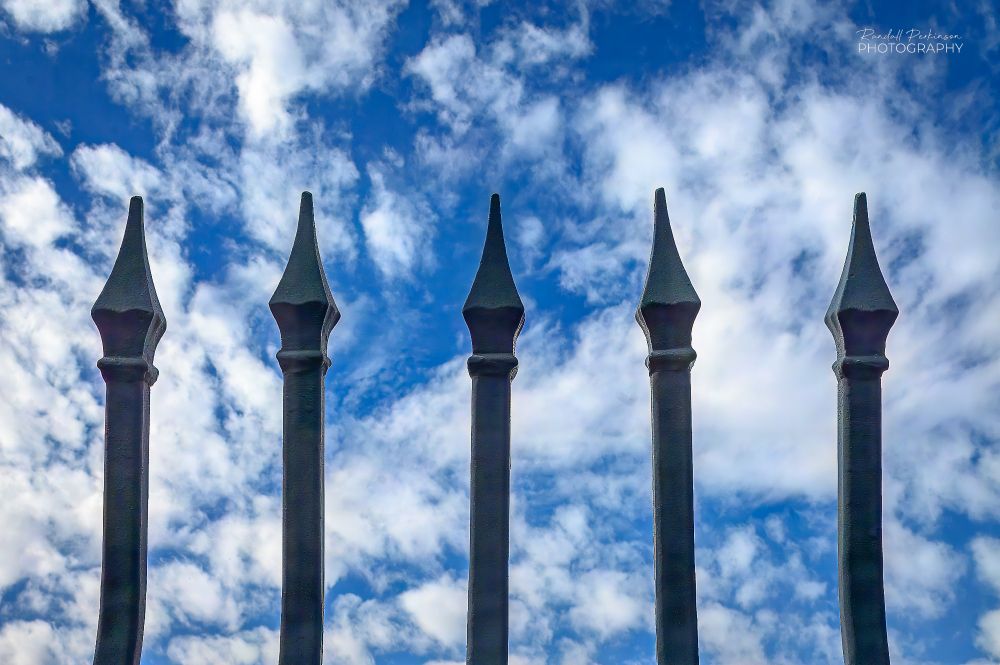 The tips of five vertical metal bridge railing member are seen in front of a blue sky with lots of white clouds.