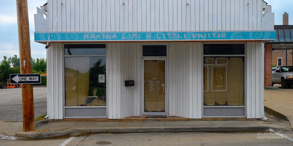 A former candy store building with a center entrance and a window on either side.  Vertical white siding covers the front of the building.  An overhang has a blue band around with the words, "Making life a little sweeter".