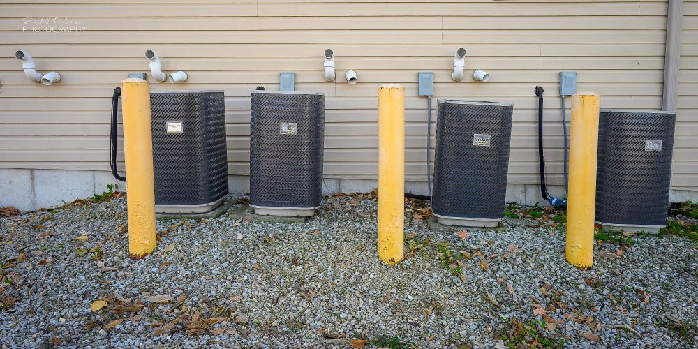 Four AC units in varying degrees of plumbness sit behind 3 yellow pipe bollards.  On the building wall behind are 4 sets of 2 vents.