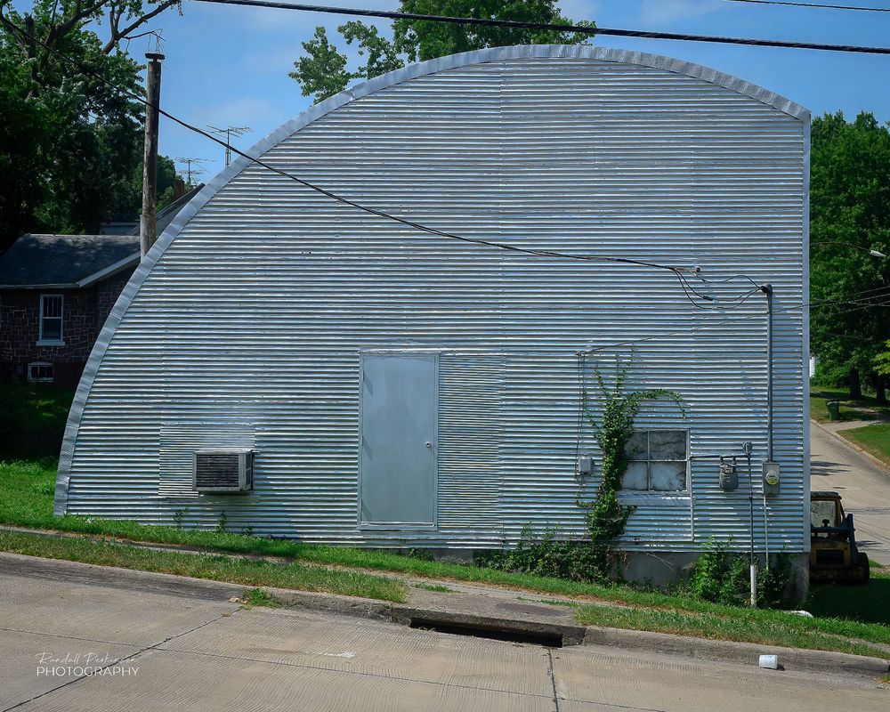 A Quonset hut with the one door, one window and an air conditioner in the end.  The right side of the arched shaped is truncated by a vertical wall.