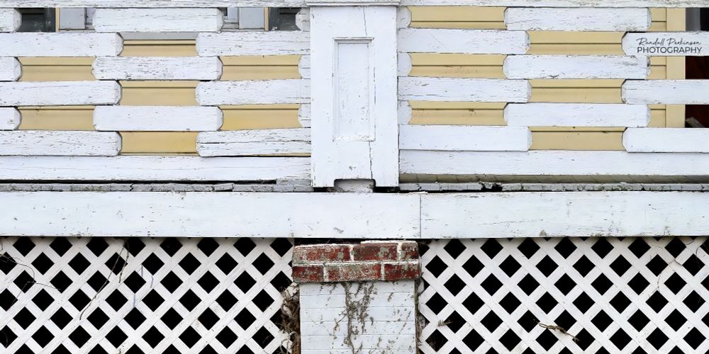 A view of a porch with white, wood lattice covering the area under the deck and a railing of stacked wooden boards about 24 inches long with about 4 inches overlapped at each end providing a contrast of diagonal and horizontal shapes. 