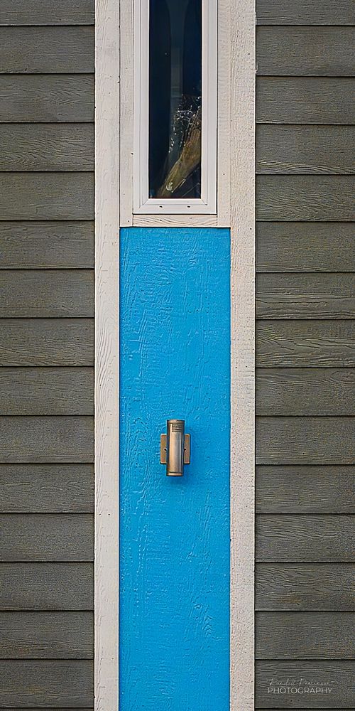 A building with grey siding has a think strip containing a blue panel with a light fixture and a tall thin window above.