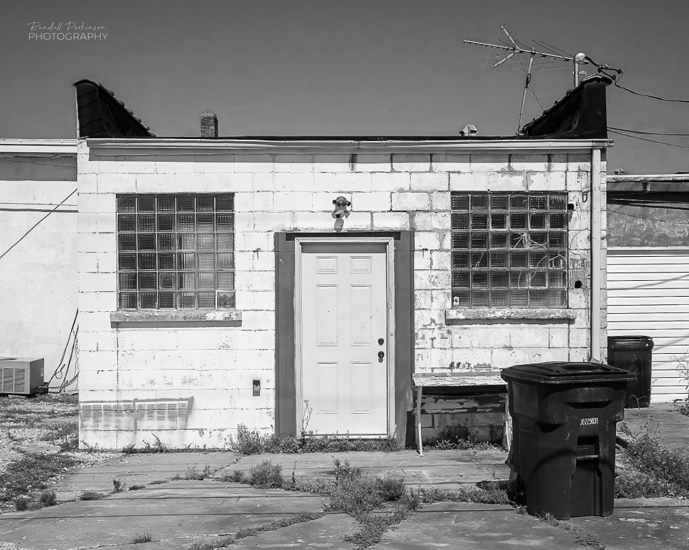 The back door of a one story concrete block building painted white.  There is a single entry door in the center and glass block windows on either side.  A trashcan sits on cracked concrete pavement with weeds growing in the cracks in front of the building.