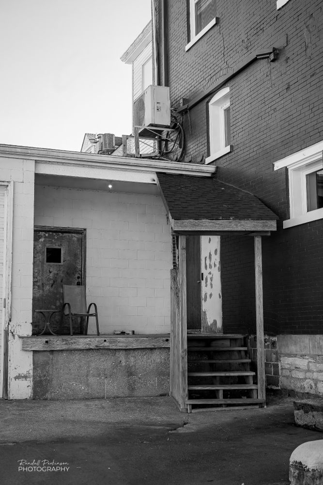 The back side of a 3 story brick building with a concrete and concrete block addition forming a small loading dock.  There is a covered wooden stairwell from the parking lot up to the loading deck area.