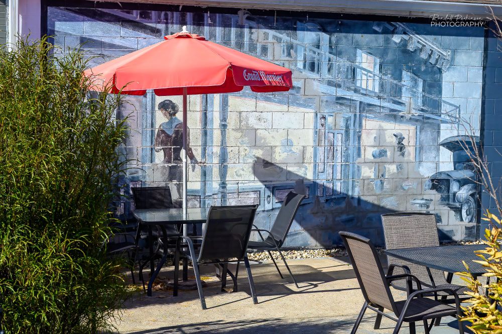 A wall painting of a street scene with 2 story buildings and a women painted in shades of blue is behind a restaurant patio with black metal chairs and tables and a red table umbrella.
