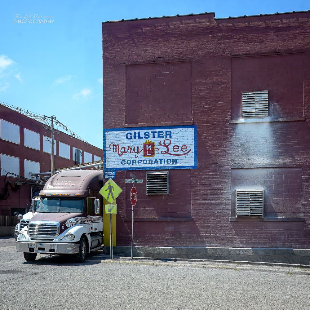 A two story brick building with a sign that says "Gilster Mary Lee Corporation" has exhaust fans in several of the bricked in windows.  The second story vent has exhausted white flour onto the wall below it.  A semi truck is parked next to the building on the left side.