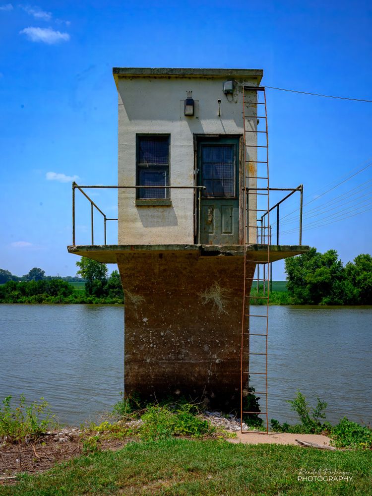 A small water intake tower rises above the shoreline of the Kaskaskia River at Evansville, Illinois. Access is via a steel ladder to a raised concrete deck that encircles the tower.  There is one door and one window at the top of the tower