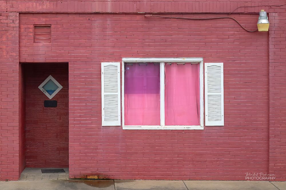 The front of a brick building painted a pinkish red has a white window with white shutters.  Inside the window are pink and purple curtains.