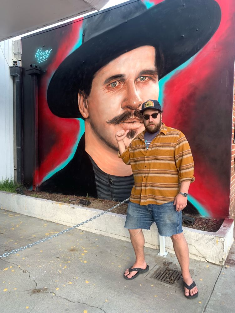 A photo of Ash in front of a mural of Val Kilmer from Tombstone in Santa Monica.