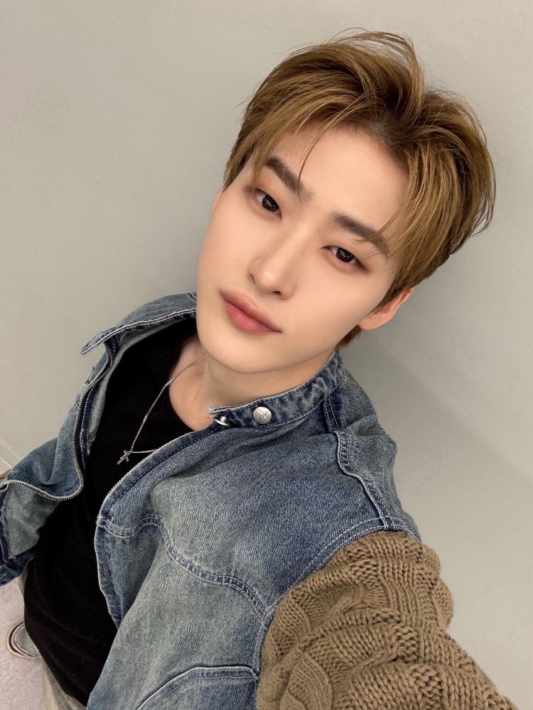 all(h)ours youmin! he has light brown / dark blond hair. he’s wearing a denim jacket with sweater sleeves over a black shirt. 