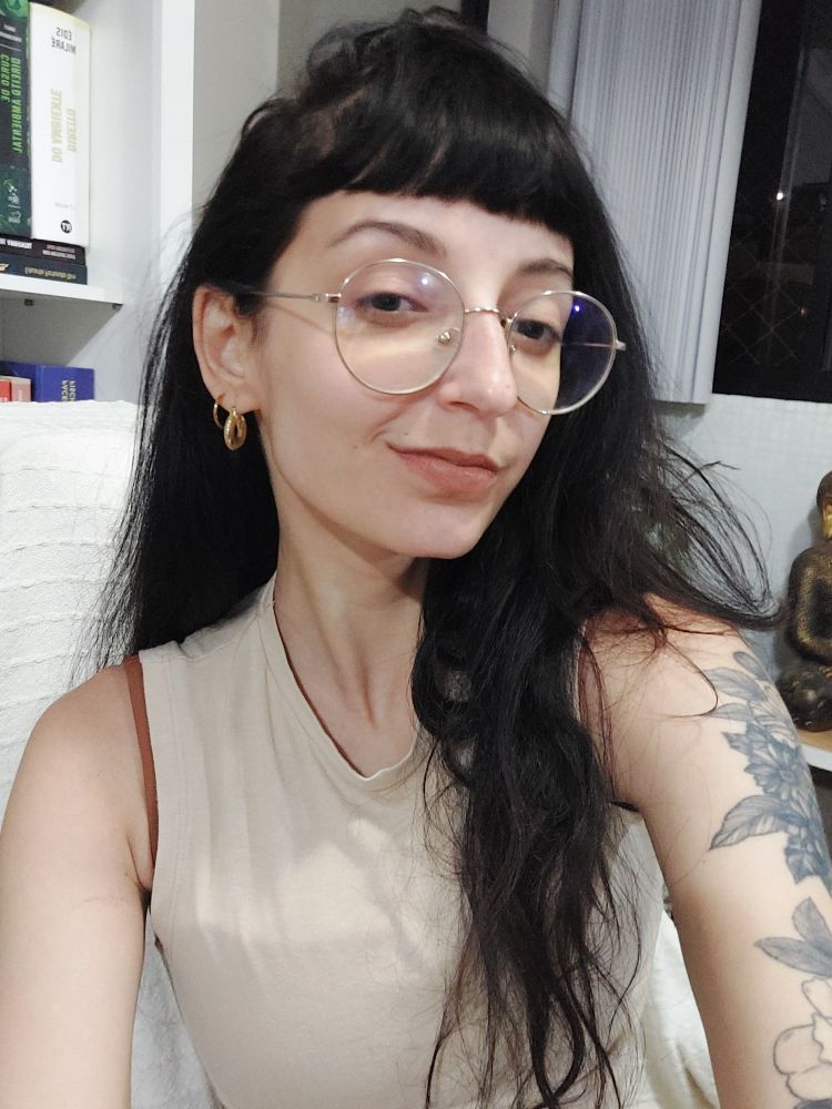 foto minha. eu sou uma mulher branca, de cabelo preto e óculos. estou olhando para a câmera e sorrindo um pouco, sem mostrar os dentes. o batom é um nude com fundo quente.