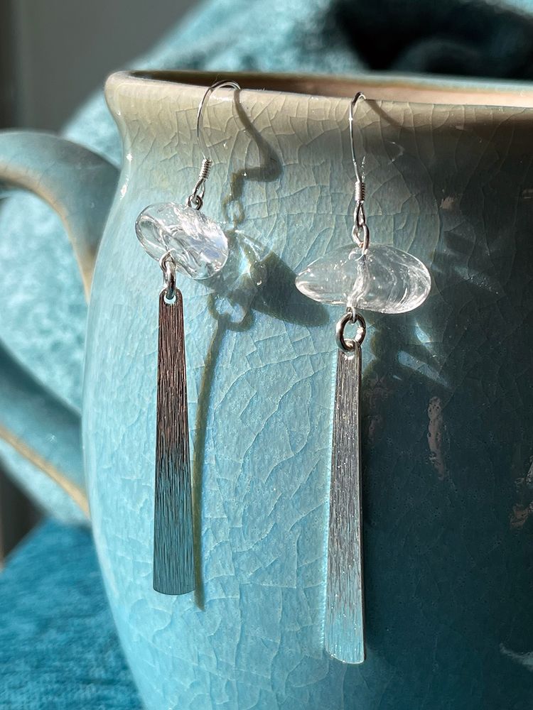 Dangle earrings featuring a textured silvertone bar charm and clear quartz pebbles, hanging on a blue ceramic teapot