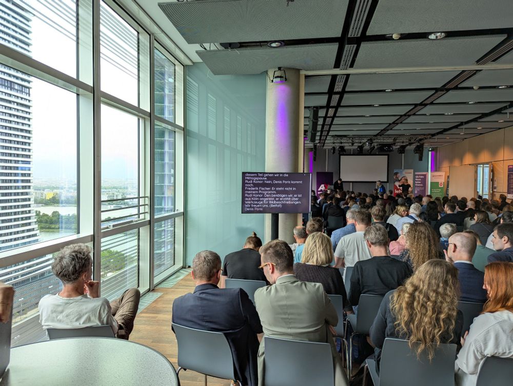 A-Tag Accessibility Conference at Sky Stage: A large audience sits facing a stage during the A-Tag accessibility conference at the Sky Stage of Tech Gate Vienna. The room features floor-to-ceiling windows, purple accent lighting, and banners along the walls. A presentation is underway with live captions shown on a screen to the side.