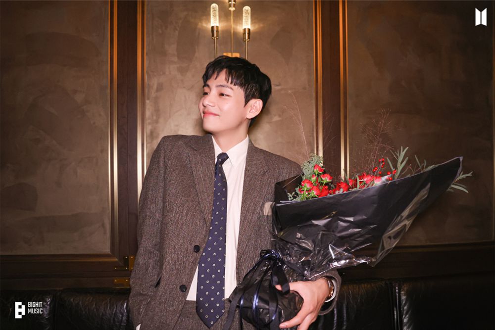 Tae in a warm brown suit carrying a big bouquet of red flowers.