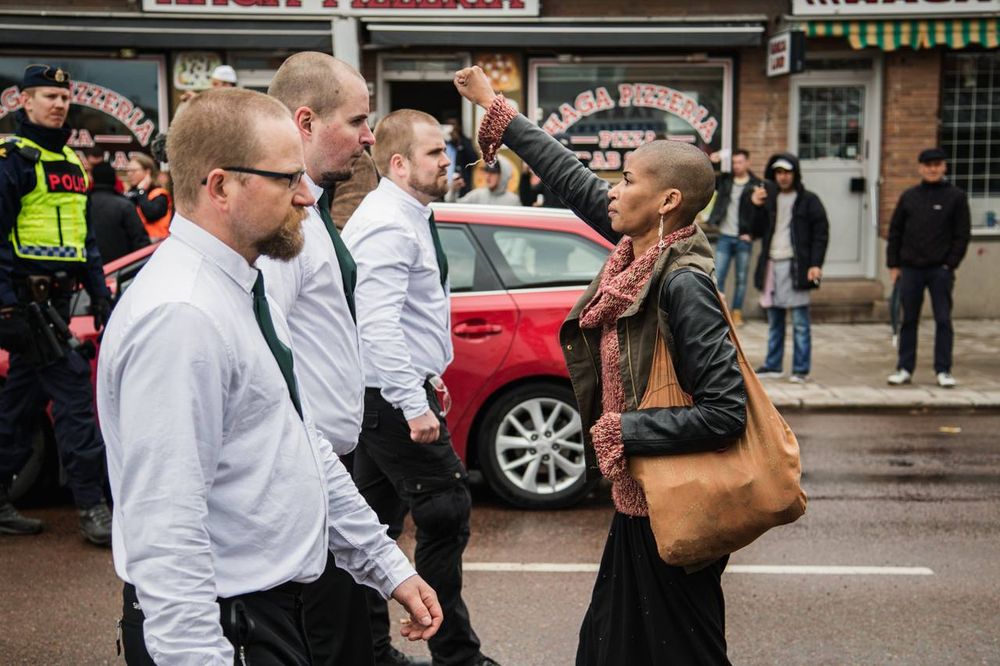 Photo montrant Tess Asplund le 1er mai 2016, s'opposant à une manifestation de néo-nazis à Borlänge, en Suède. Elle fait face à 3 néo-nazis, au milieu de la rue, le poing levé et l'air calme et déterminée.