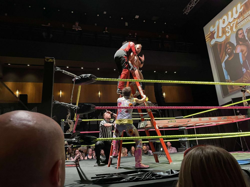 A wrestling ring with Stuart the referee looking flabbergast as MRB holds TY Jackson at the top of a ladder while Stephen Crowe in a fancy cowboy outfit steadies it