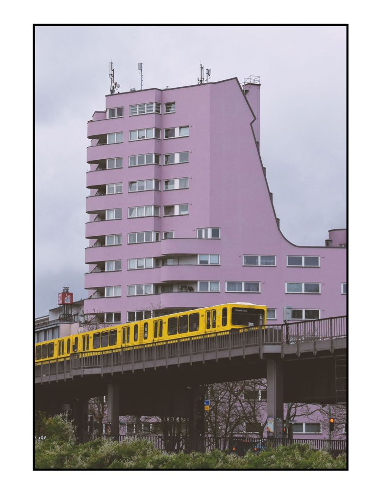Photograph of a yellow metro train travelling on a raised track. The train is passing a pink coloured tower block. The sky is grey.