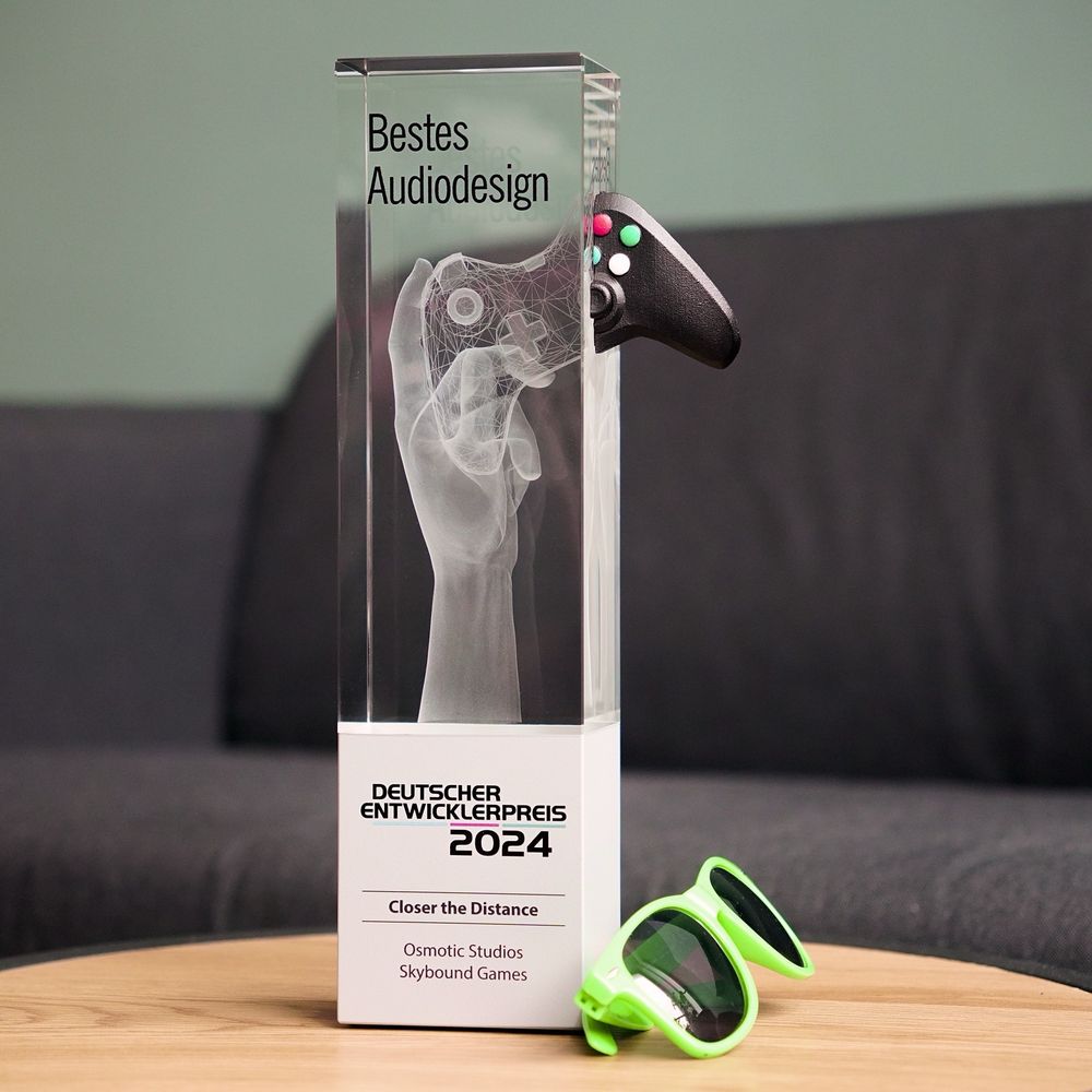 The german developer award 2024, made out of metal and glass. In the inside, there's a hand, holding a game controller. And next to the award, there are green sunglasses.