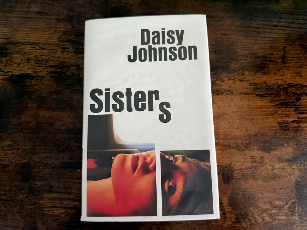Photo of a book on a wooden counter. 
The book is ‘Sisters’ by Daisy Johnson and has a white cover with a photo from the neck up of a girl laying down looking to the camera. 